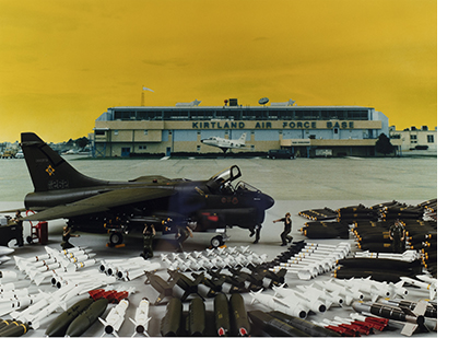 a digital composite photograph of a fighterjet surrounded but orderly lined up bomber. In the background a building says Kirtland Air Force Base The sky is solid mustard yellow