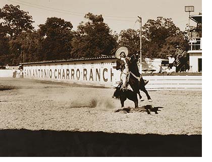 A black and white photo of a man riding a bucking horse in an outdoor stadium a wooden wall in the background says San Antonio Charro Ranch 