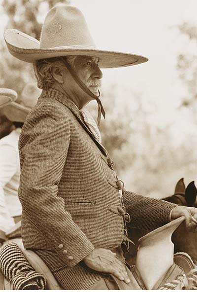 A black and white photo of a man sitting straight backed and proud on a horse he is wearing  a tweed coat and large sombrero
