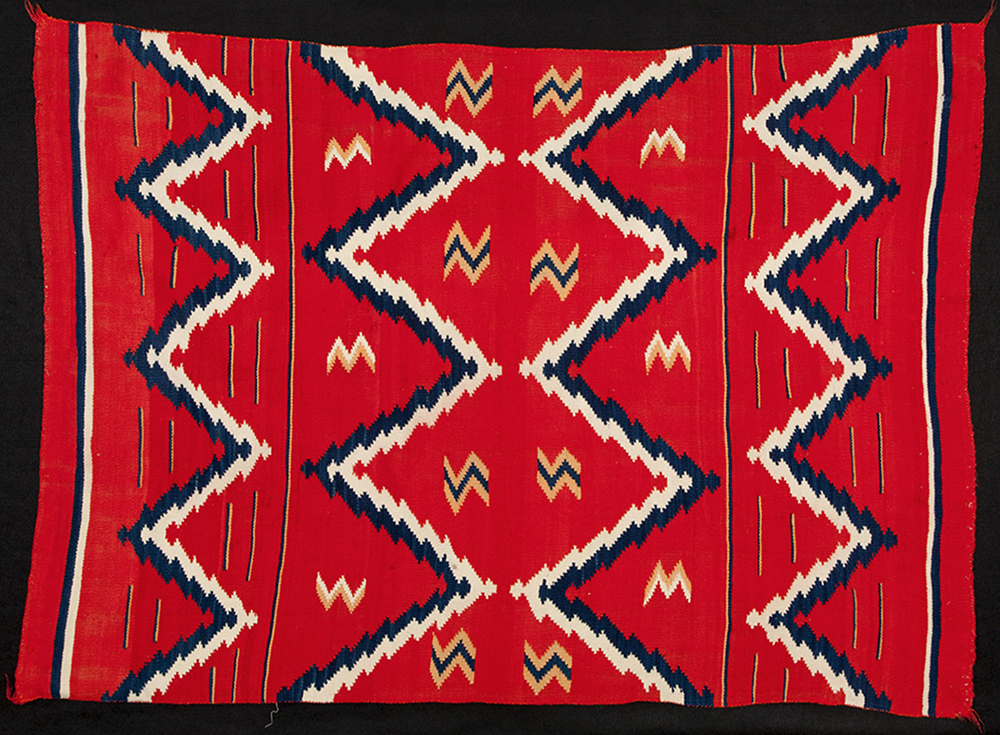 A predominantly red woven rug with four blue and white jagged zig-zag lines running horizontally across the rug. Smaller zig-zag shapes are dispersed around the middle area between the lines. Thinner and shorter blue and white lines run between the larger zig-zag lines on the top and bottom of the rug. 