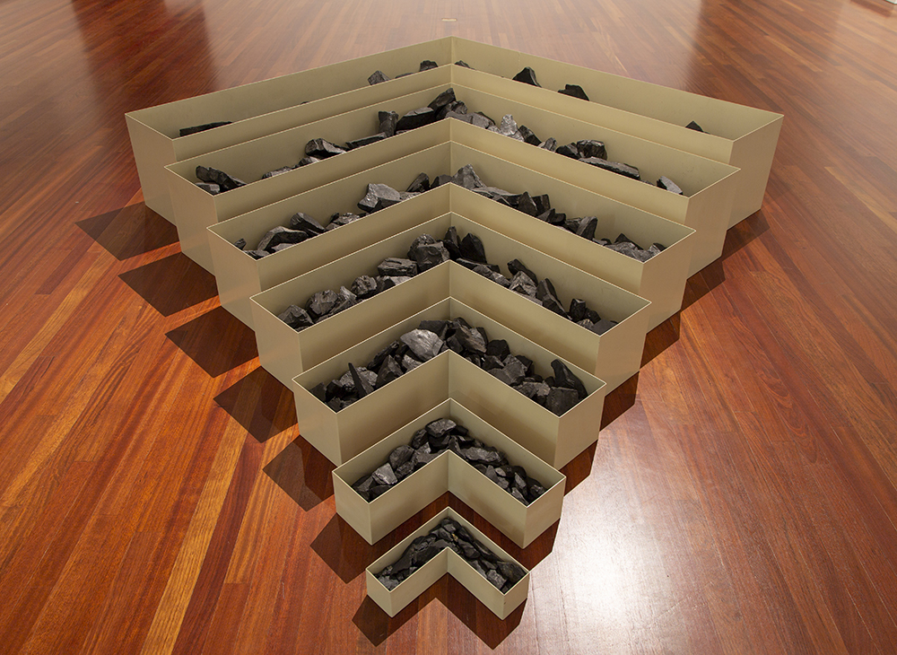 A sculpture of L-shaped buff-colored metal boxes are nestled together on the floor forming an arrow shape. The seven boxes gradually increase in size and contain chunks of black coal.  