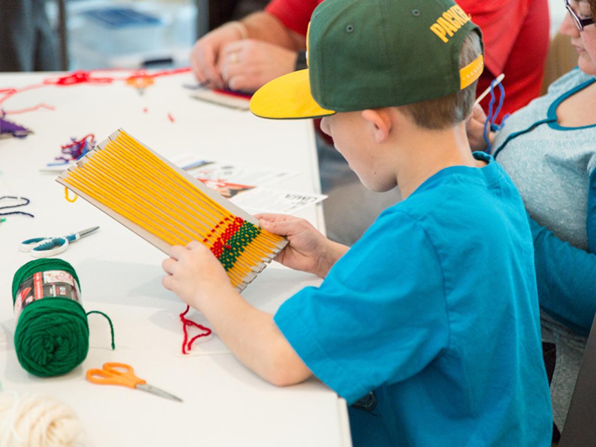 Boy in blue shirt and green and yellow baseball hat weaving a matching opt holder on cardboard loom