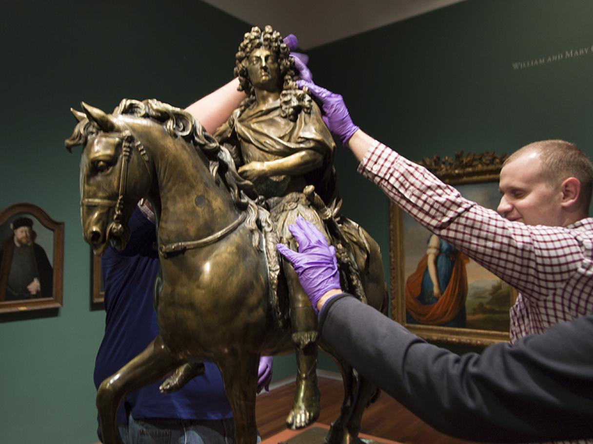 Three people reach up to touch statue of King Louis on horse back all are wearing purple nitrile gloves