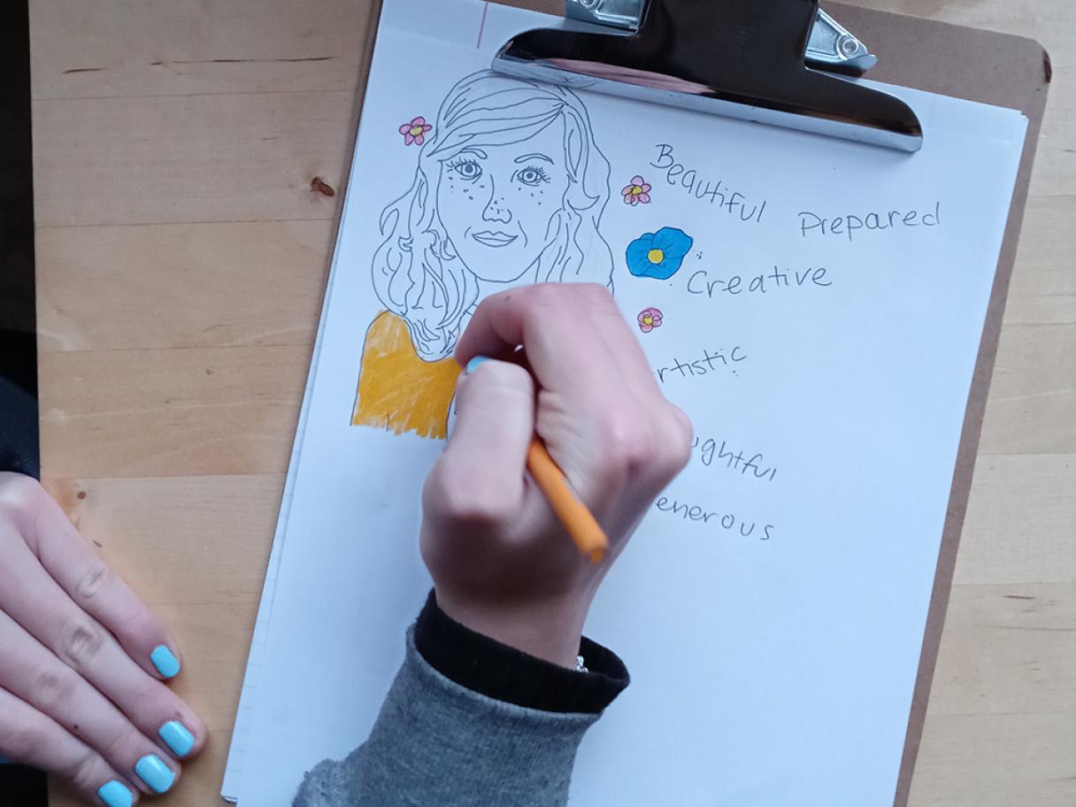a child's hand holds a yellow pencil drawing on a clipboard. A white piece of paper has a drawing of a black outline of a girl only the shirt is colored yellow