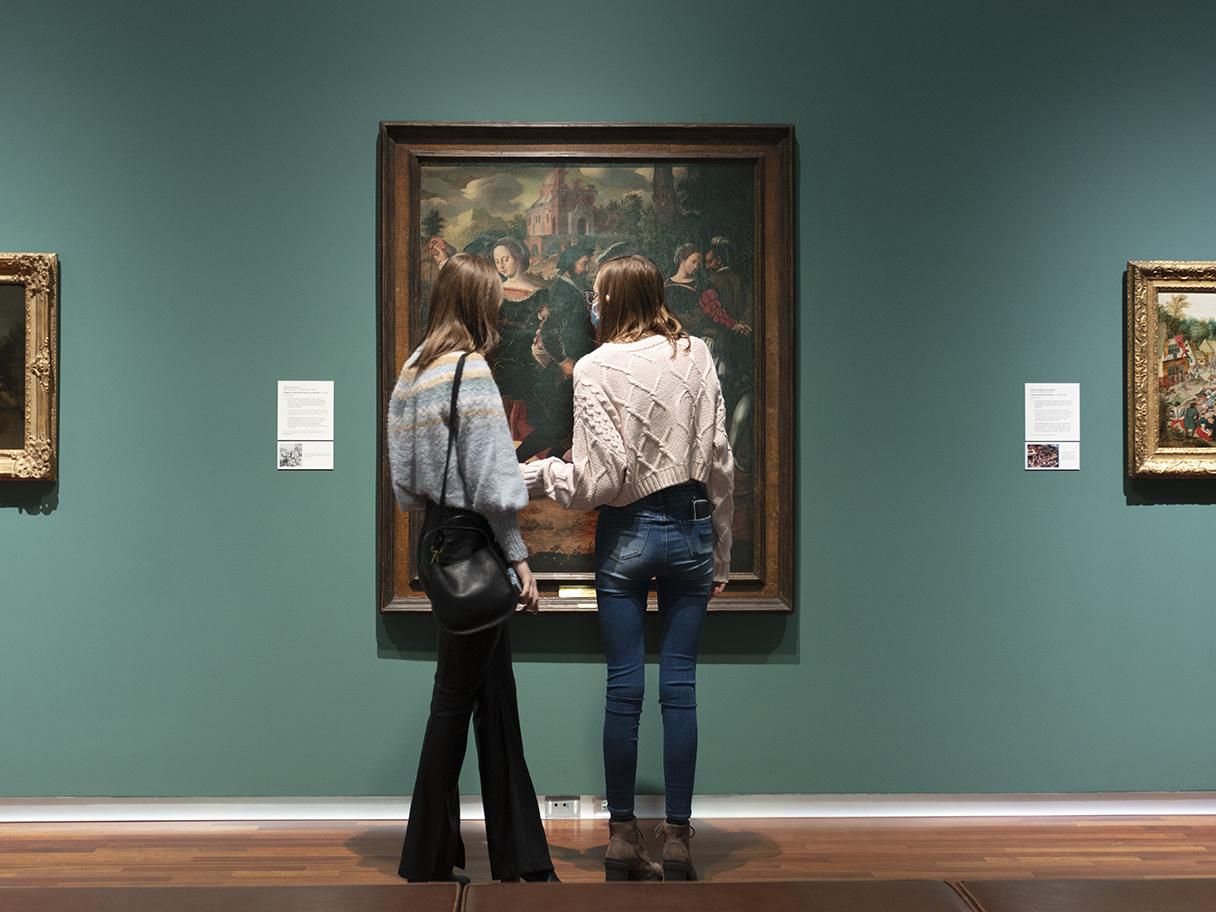 Two women stand in front of a large oil painting conversing in the UMFA's European gallery.