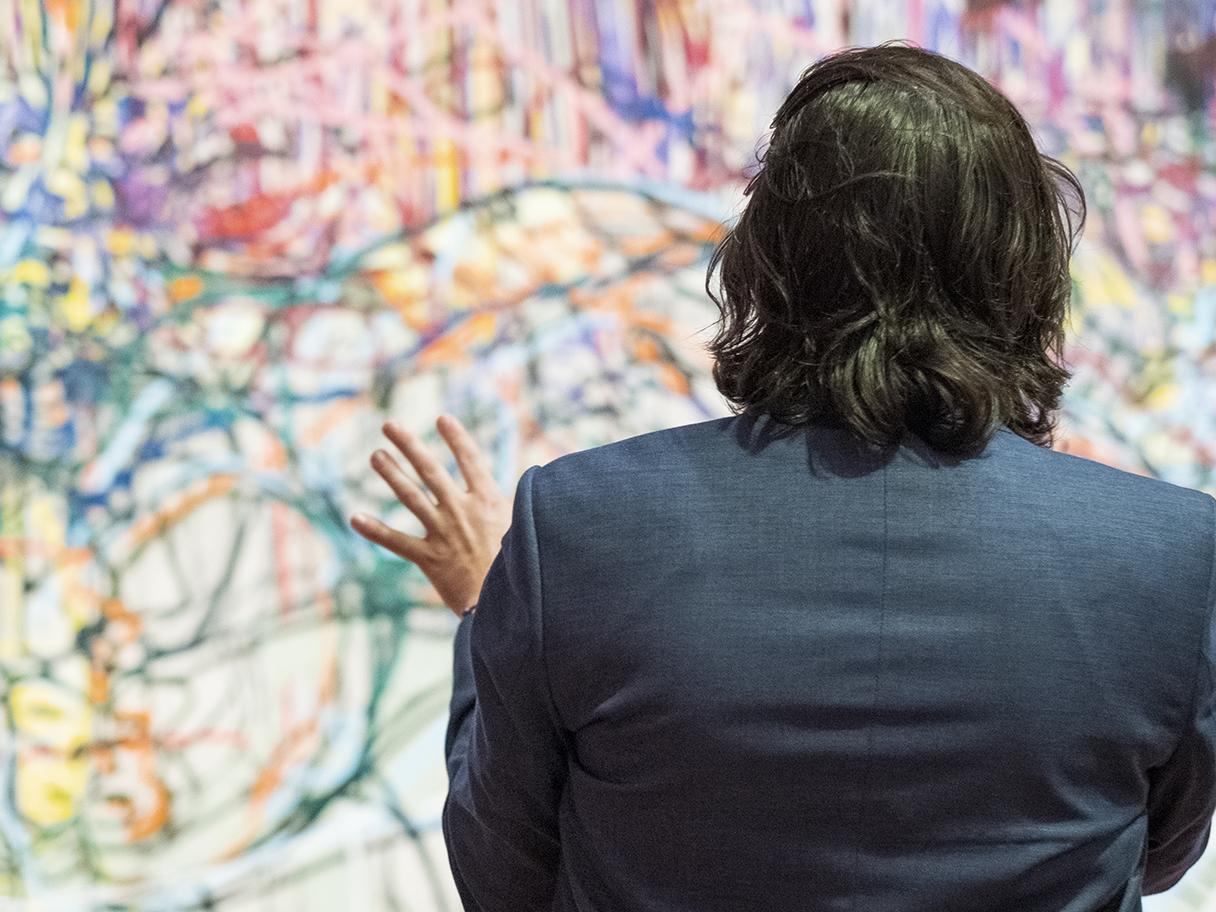 A man in a blue suit with shoulder length hair, looking at a large abstract painting. 