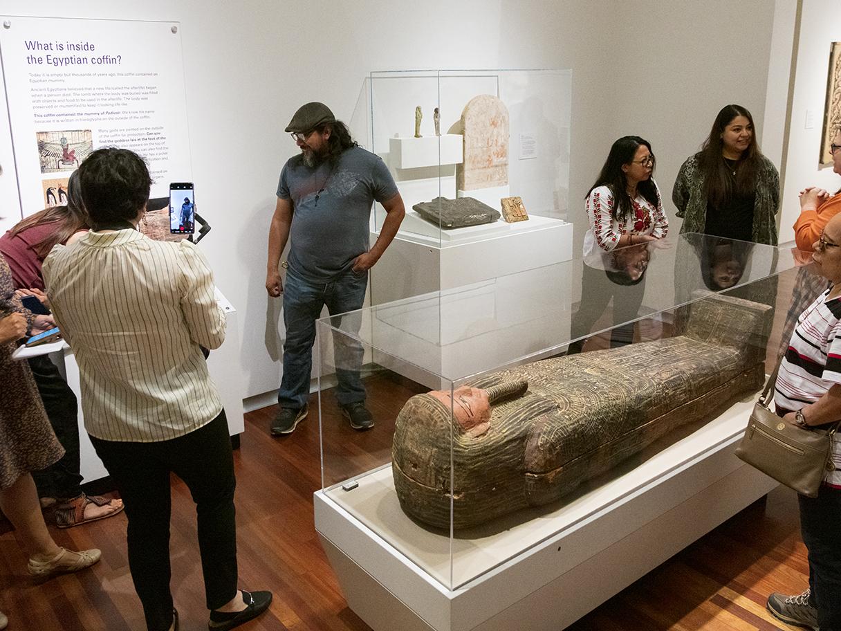 A group of people stand around a white room, looking at different artifacts in display cases. There's a mummy in the main display case in the middle of the room.