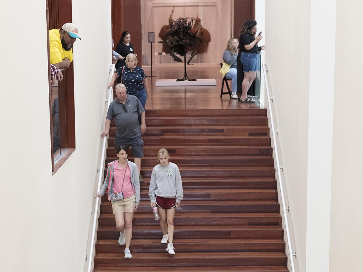Museum visitors walking down the main stair case, a man in a yellow shirt and baseball cap looks down at the stairs from a window on the left.  