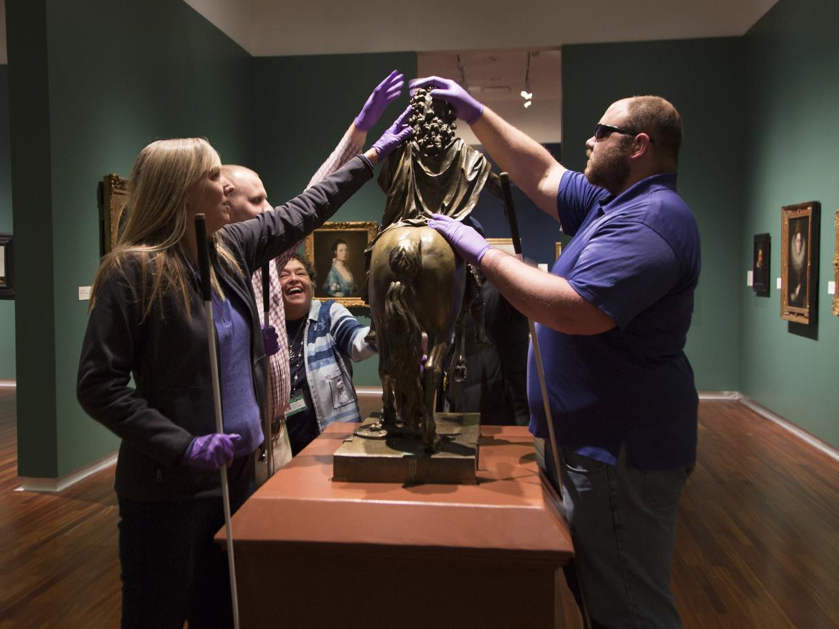 Image of a touch tour in the European gallery. The seeing impaired guests touch a bronze sculpture, wearing purple gloves to protect the art. 