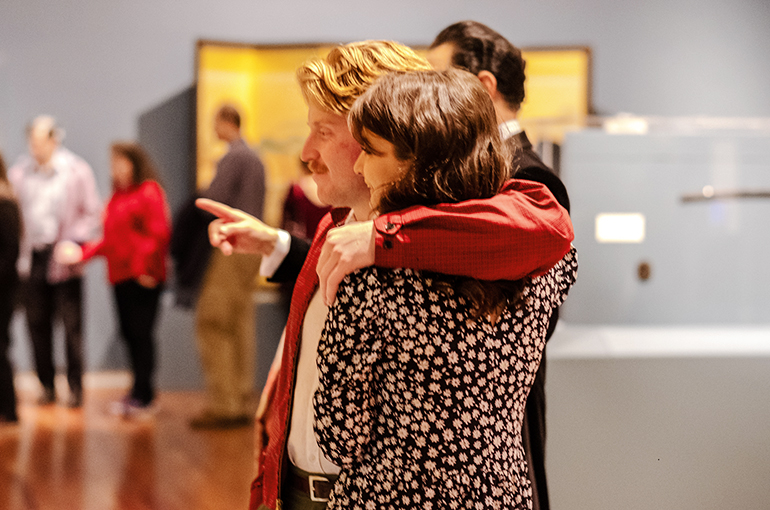 Couple at UMFA Valentines Evening 2020 A couple view Beyond the Divide at the UMFA Valentines event, man stands with arm around woman looking off towar art out of view.