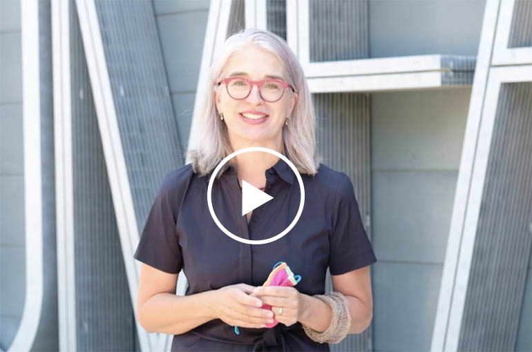 Gretchen Video play button Gretchen Dietrich in front of UMFA building she wears a navy blue short sleeve shirt dress and pink glasses.