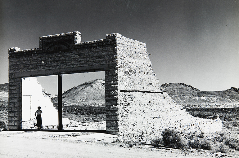 a vintage balck and white photo of a ruined rock building in a dessert, there is a woman standing in a large square opening the looks like it could be a garage door