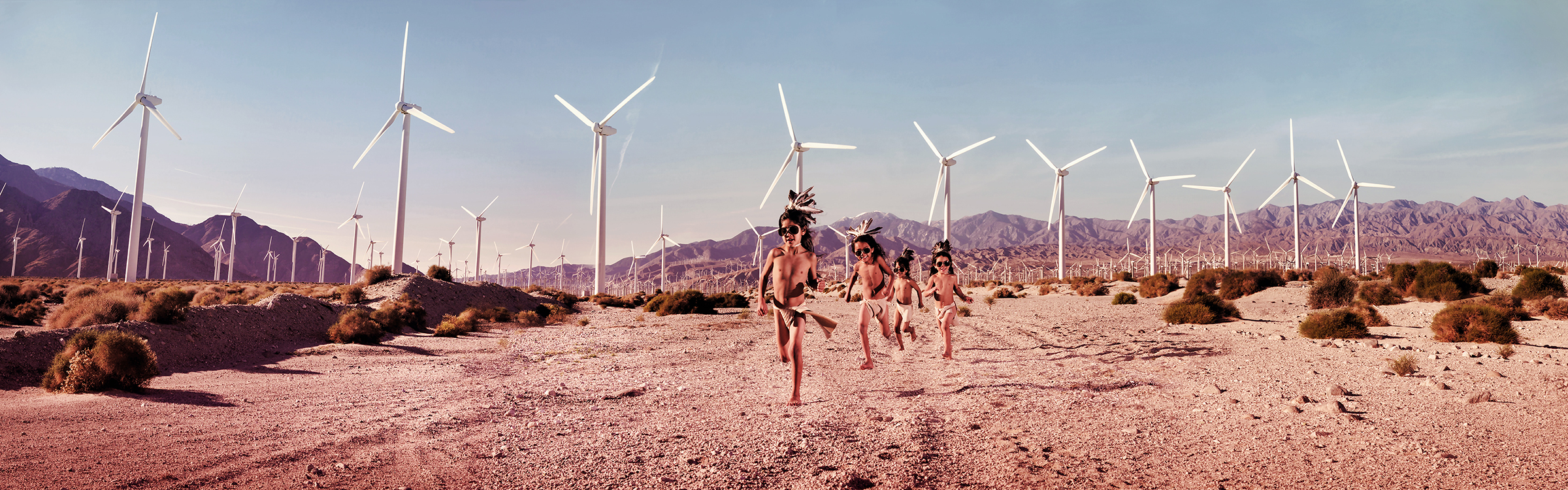 four children in indigenous dress run joyfully in  a dessert field with  wind turbines 