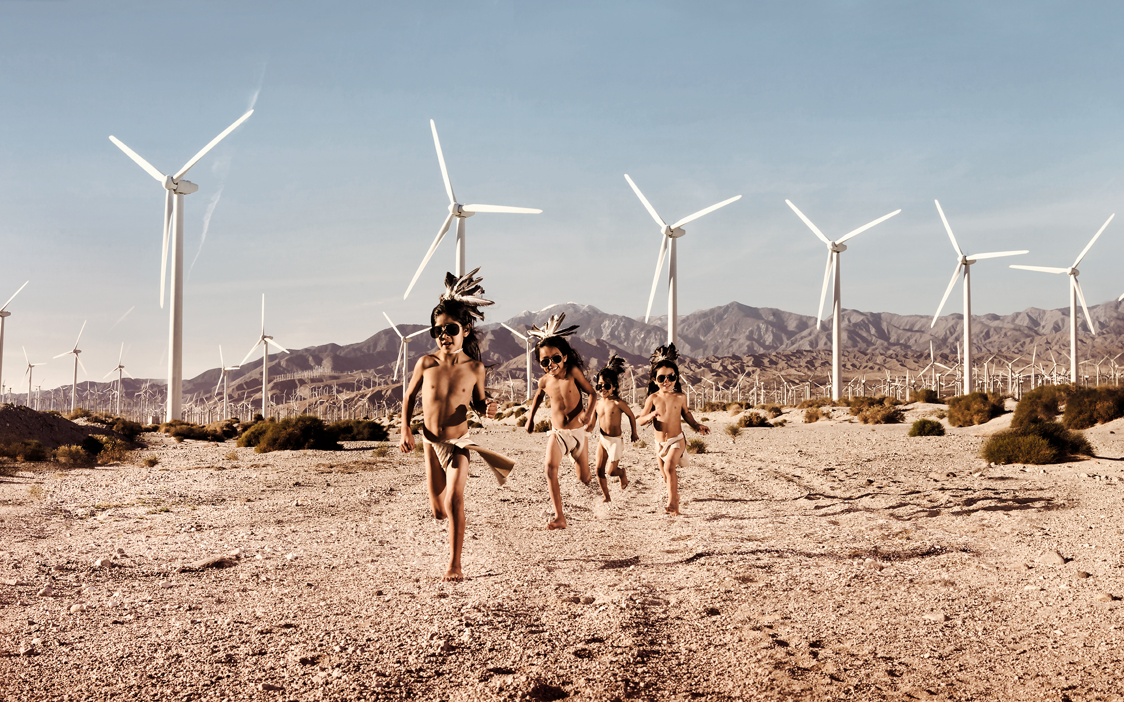 Four indigenous children in sunglasses and traditional clothes  run across a dessert field with wind turbines behind them