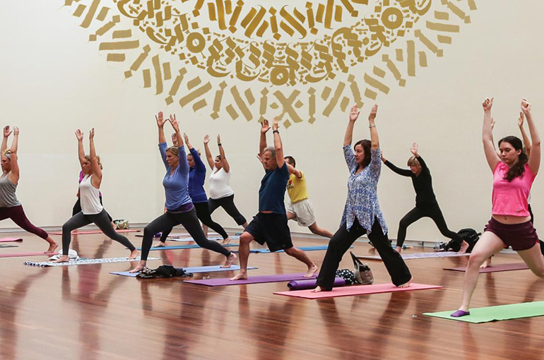 a group of twenty people stand in in warrior pose during a yoga class in the UMFA Great Hall