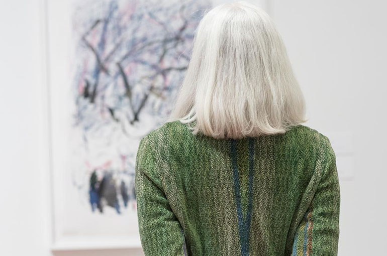 A women with shoulder length gray hair wearing a green sweater stands with back to the camera looking at a an abstract painting.
