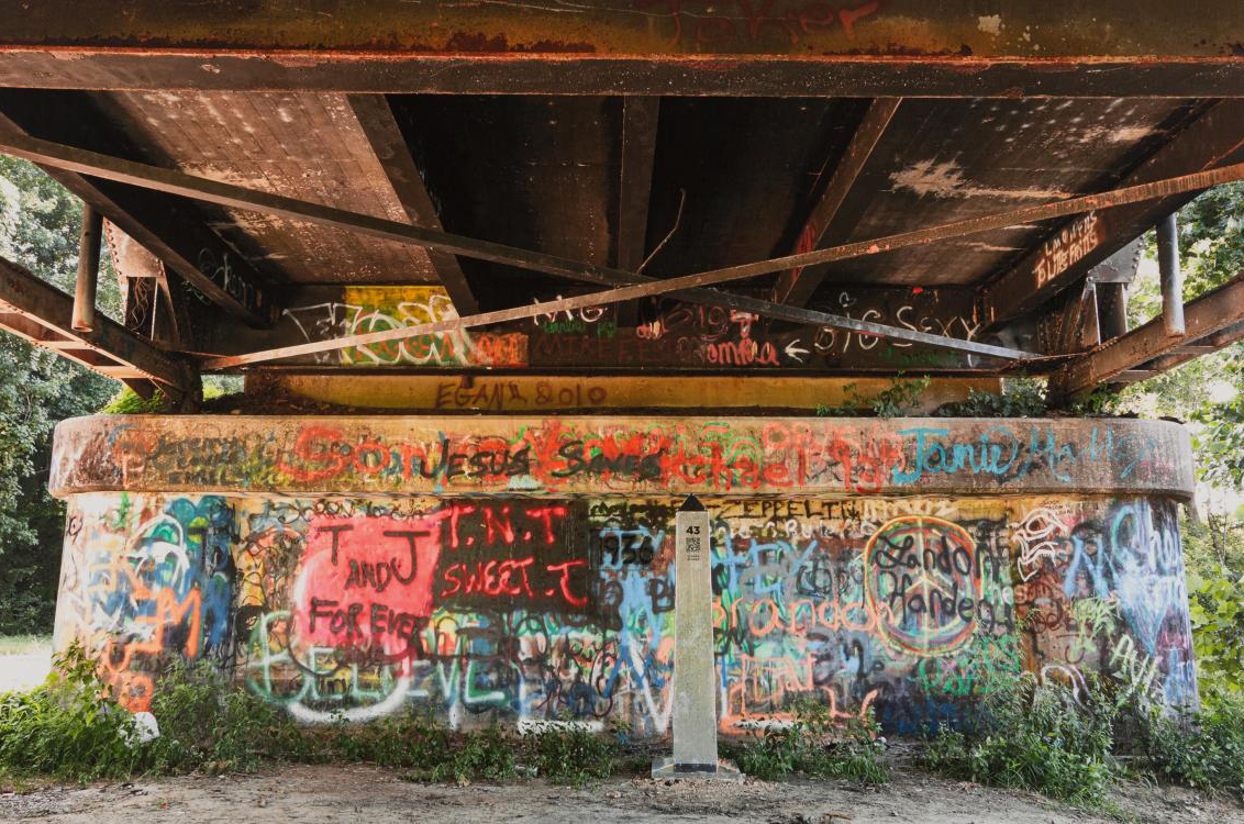photo of an underpass with a concrete wall covered in graffiti  there is a large metal obelisk about 6 feet tall in front of the wall