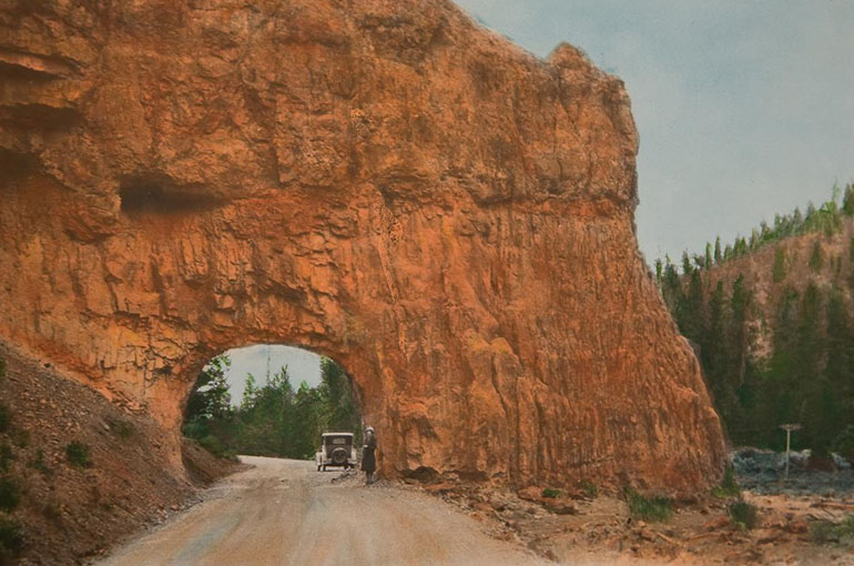 an antique hand colored photograph of a red rock wall wtih a tunnel going through it there is a woman standing next to a car in the tunnel