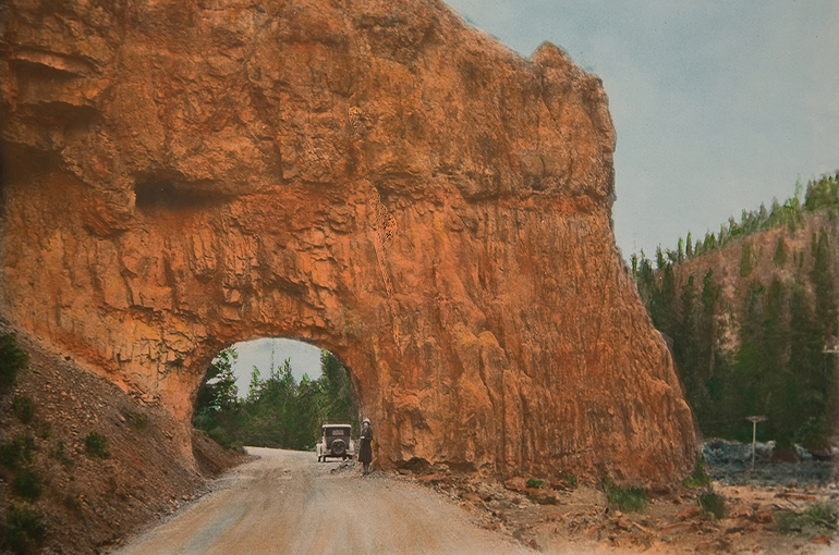 A road runs through a large, red rock cliff. There's an old car on the road and a woman walking behind the car. There are trees and a blue sky in the background.
