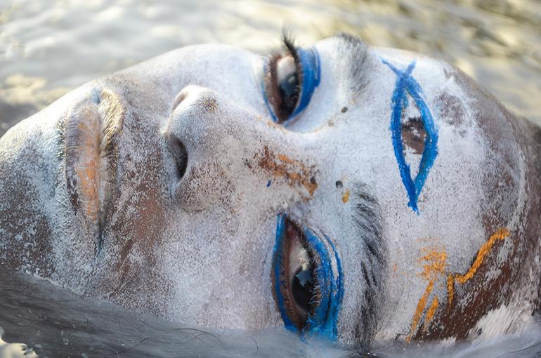 A black woman with a white painted face, blue paint on her eyeballs and an eye painted on her forehead, lays with her head half way under water. She is looking directly at the camera.