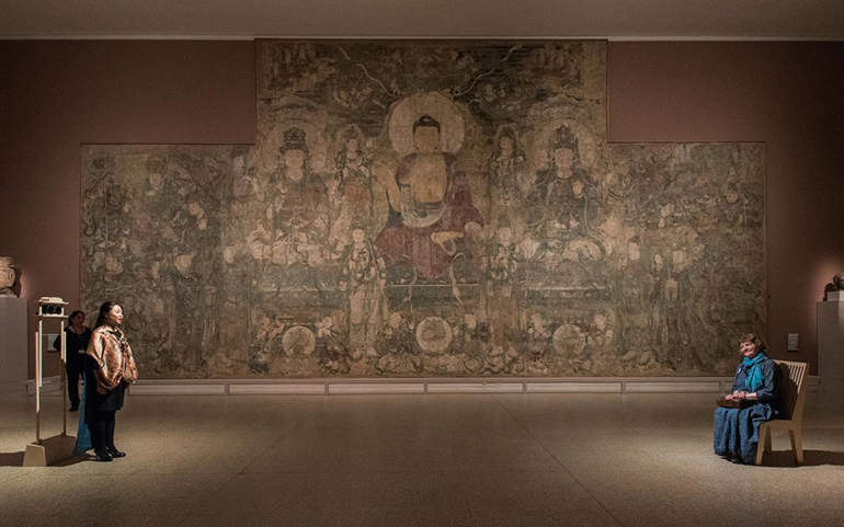 A woman stands on one end of a long room and another person stands on the opposite side, facing her. They are standing in front of a large mural of deities in gold, green and red.
