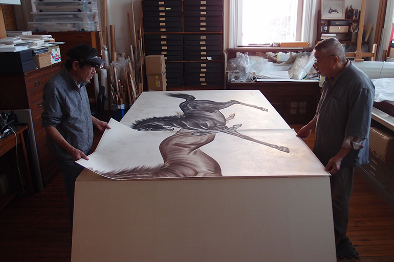 Two men stand on opposites sides of a wide, tall table. There's a drawing on two large pieces of cream paper with a brown horse on one and a black horse on the other. The men are handling and lifting the paper.