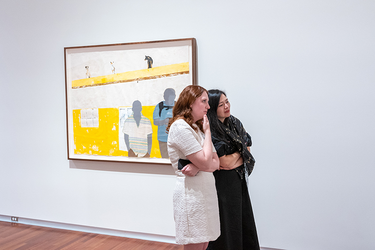 Two women in dresses stand looking at something out of view. Theres a yellow framed artwork of two human silhouettes in the background hanging on the wall.