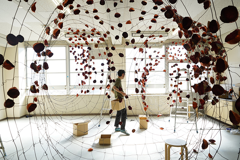 A man stands in the middle of a room with boxes, stools and ladder and small rock shapes copper pieces hanging from wire spread across the room.