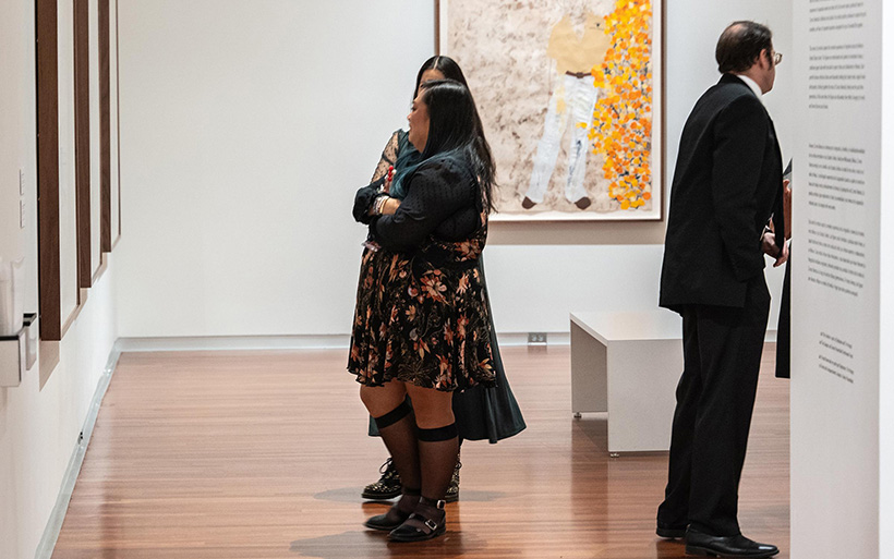 Two women with long dark hair stand next to one another, speaking and looking at art hung on a wall.