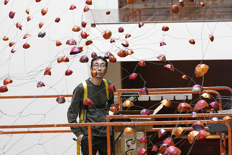 A man stands on a lift in the middle of an art installation of wires and foil, copper cups.