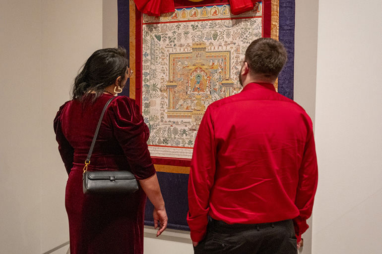 Two people dressed in fancy red clothing stand facing a gold, red and purple Himalayan tapestry.