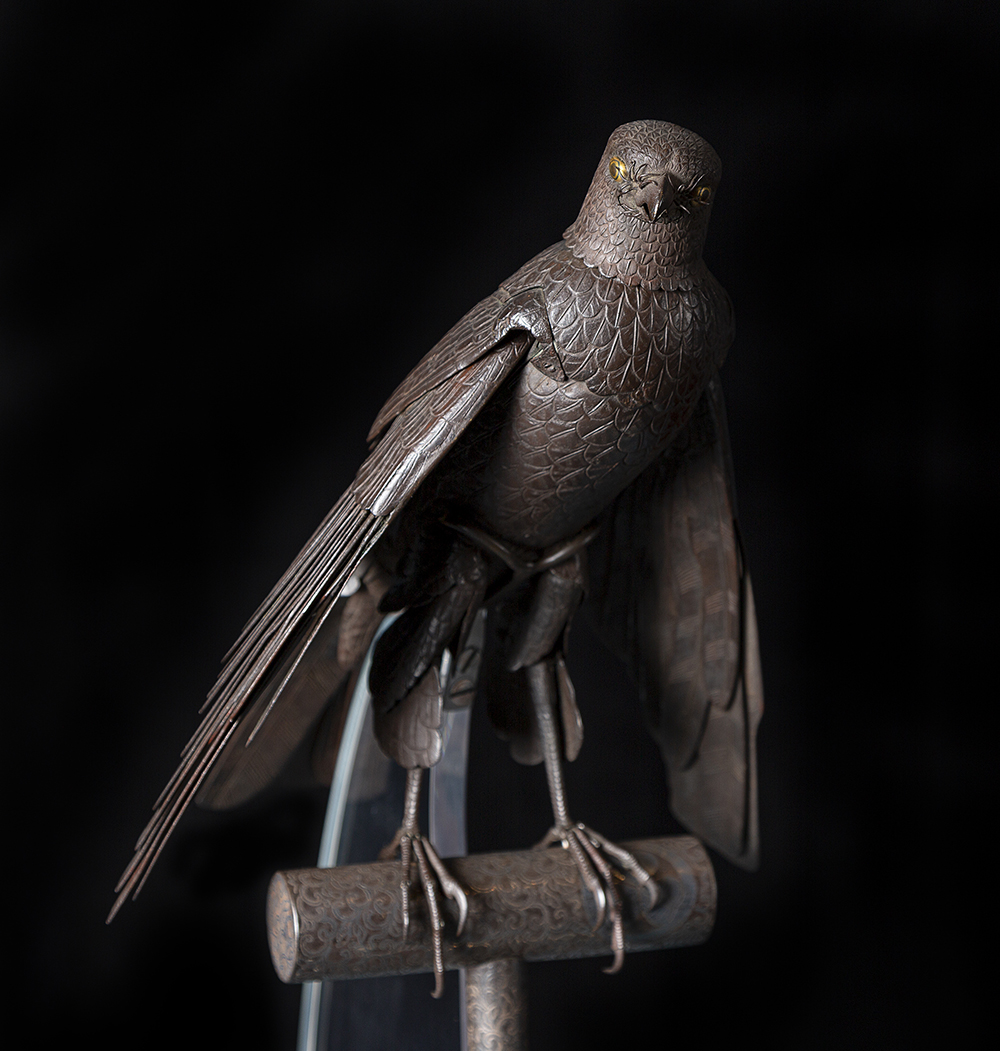 a steel sculpture of a raptor bird. The steel is a dark gray. The realistic bird has articulated wings that can move and spread out. The bird is standing on his claws on a t-shaped pedestal. 
