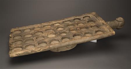 A wooden rectangular gameboard lies on a table. It has 32 circular depressions on the surface set in a grid formation. There is a handle shaped like a stick with a knob sticking out of one of the short sides.