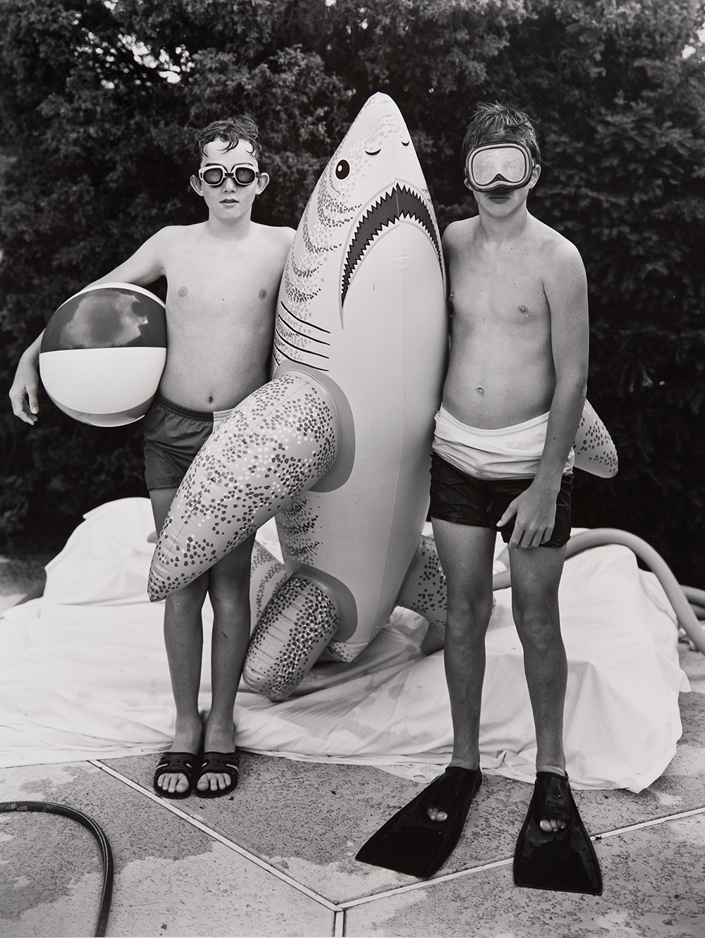 a black and white photograph of two boys standing on a pool deck. There is a blow-up shark between them and one boy is holding a beach ball. The boy on the left has swim goggles on and the boy on the right wears a swimming mask and flippers.