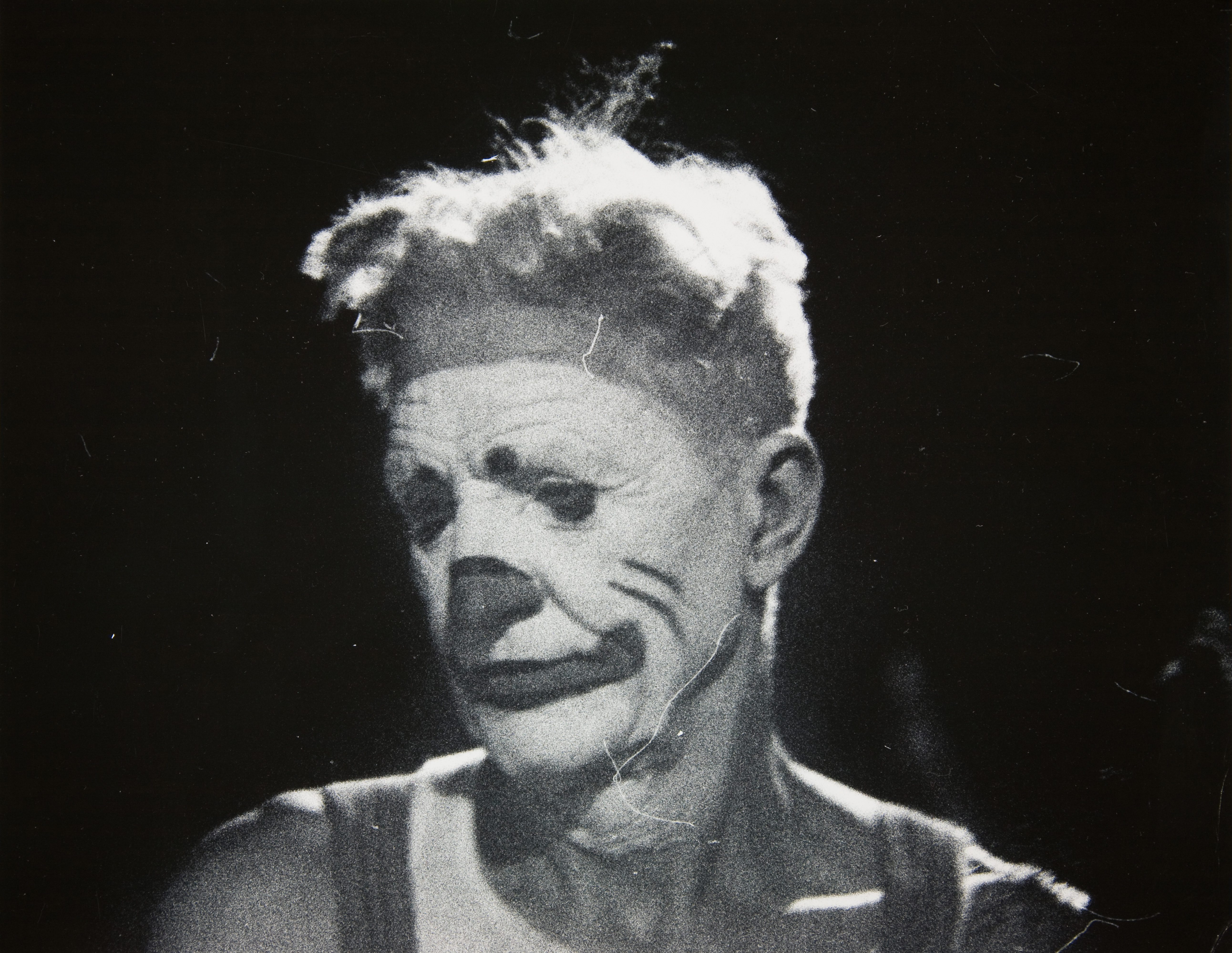 A black and white photo of figure in clown make up. The figure looks down and to the side as though sad.