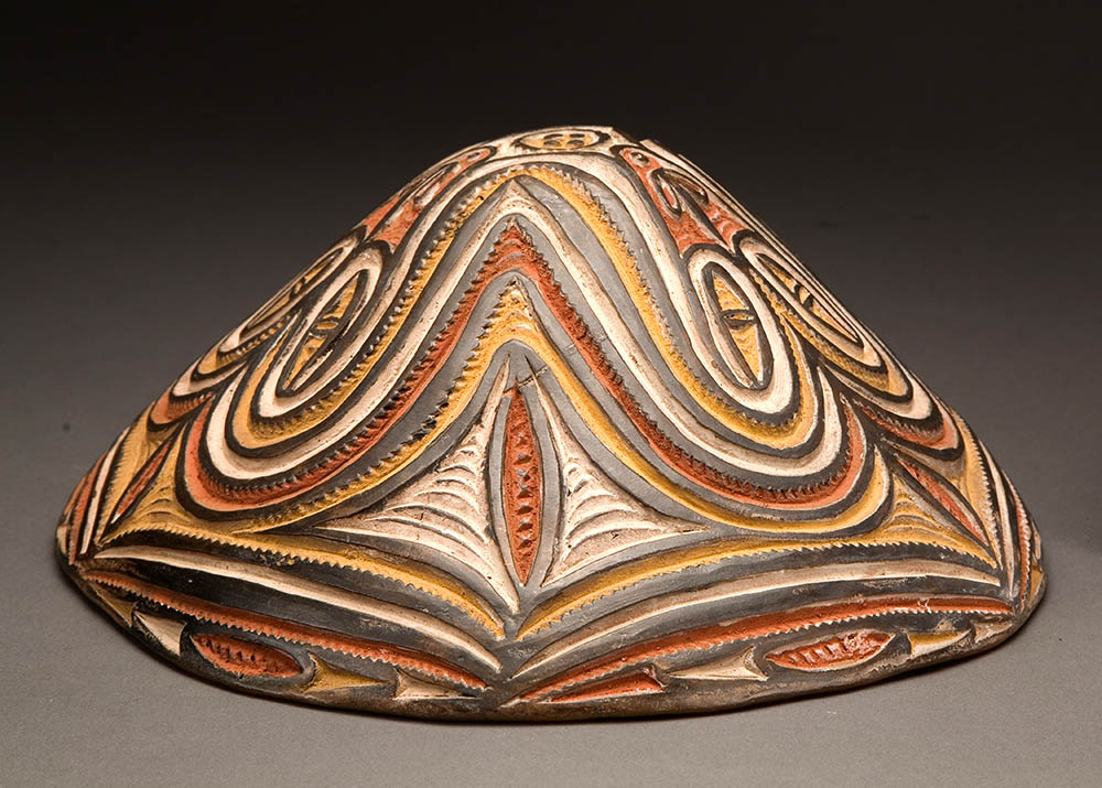 a wide conical ceramic bowl sits upside down with the bottom facing up. The outside of the bowl has incised swirled designs of lines and ovoid shapes. The raised lines are a gray color and the carved spaces between alternate colors of rust, gold, and white. 