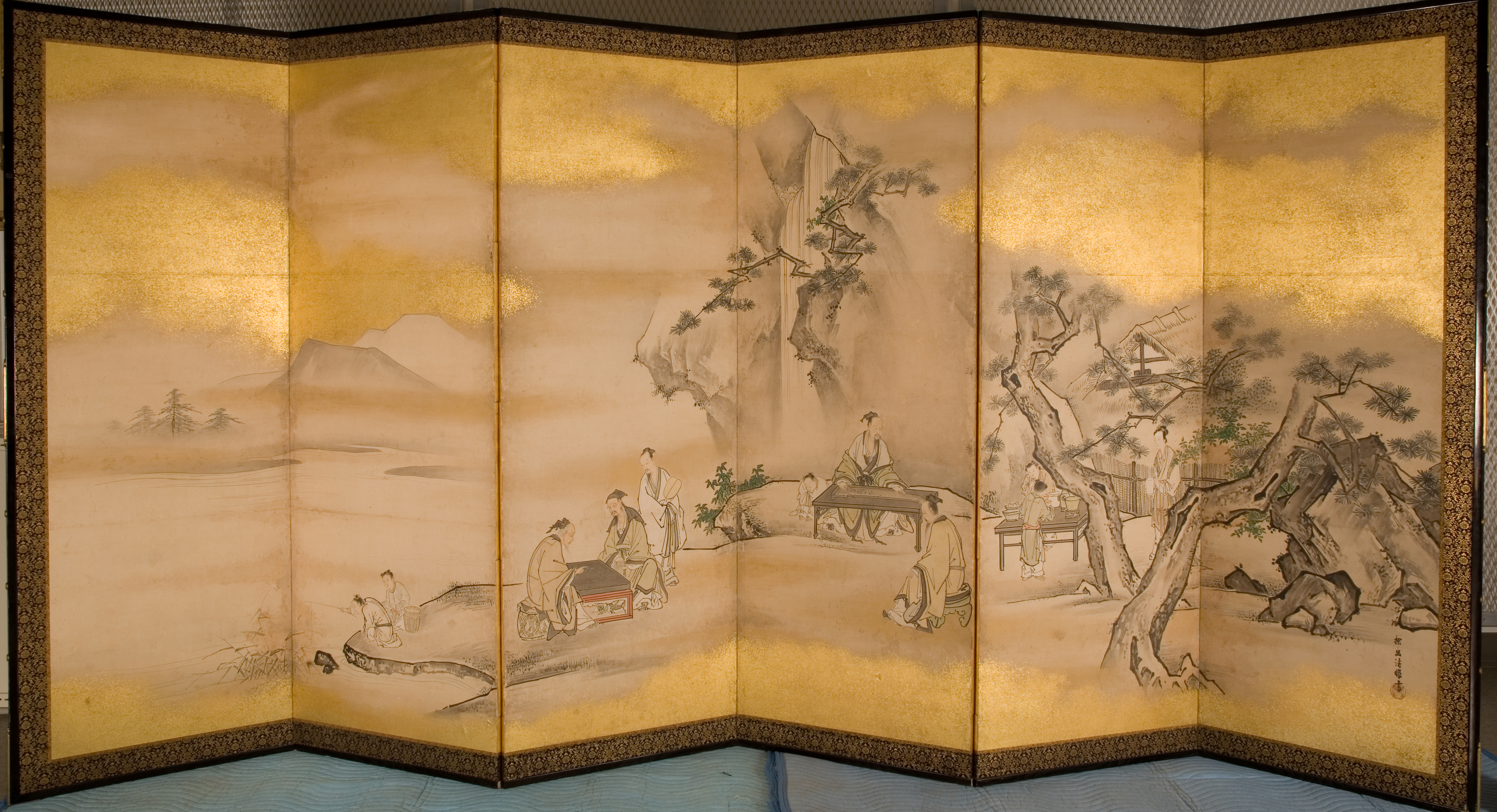 A folding paper screen with six panels depicts a landscape with gold clouds surrounding the scene. A mountain is in the distance on the upper left with a grove a trees in the lower right. Groups of people in traditional Japanese clothes are in the grove of trees doing various activities: fishing, playing the zither, painting, and playing the game Go. 
