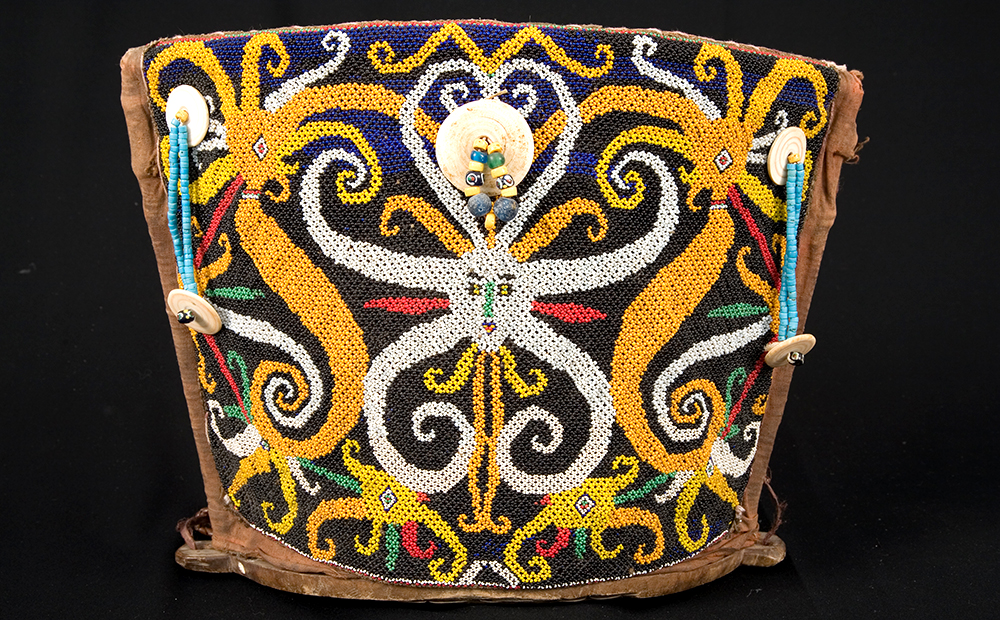the trapezoidal back of a backpack-like object. The back is covered in a beaded design of creatures with faces and octopus-like tentacles swirling in a bi-lateral symmetrical pattern. There are two string of small turquoise beads capped with a larger round shell bead on the upper left and upper right of the backpack. Attached on the upper center is large shell bead with two smaller beaded strings coming out the middle.