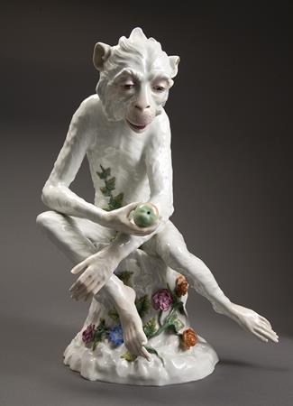 A white porcelain monkey seated on a flower stump. The monkey is holding a green apple and has vines growing up its stomach.