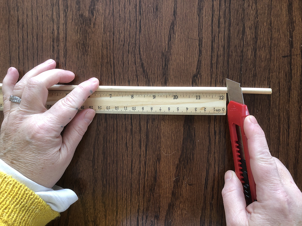 A person's hand measuring the a dowel and cutting with a razor knife