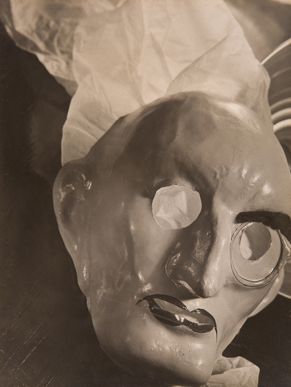 a black and white photograph of a realistic mask sits atop some tissue paper. The mask is of a bald man with a monocle on his left eye. His lips are outlined in a darker paint. 