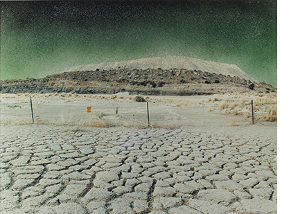 a photo of cracking gray desert sand in the foreground leading to a wire fence with a barren hill in the background.
