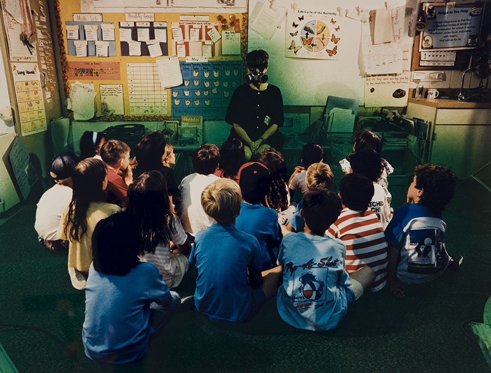 a color photograph of a teacher in a classroom with children sitting around on the floor in front of her. The teacher is wearing a gas mask. There is a green haze all around the children and the teacher.  
