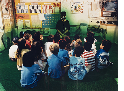 A photo of a classroom with teacher sitting on a chair with students surrounding them on the floor. The teacher is wearing a gas mask and there is a green haze around everyone. 