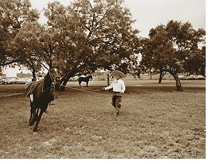 A black and white photo of a man wearing traditional Mexican clothes and a sombrero stands in a fields with a horse on a lead there are trees and another horse in the background. 