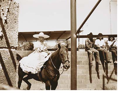 A black and white photo of a women dressed in traditional Mexican rodeo dress with a large sombrero she sits on a horse three people sit on a cinder block wall to the right 