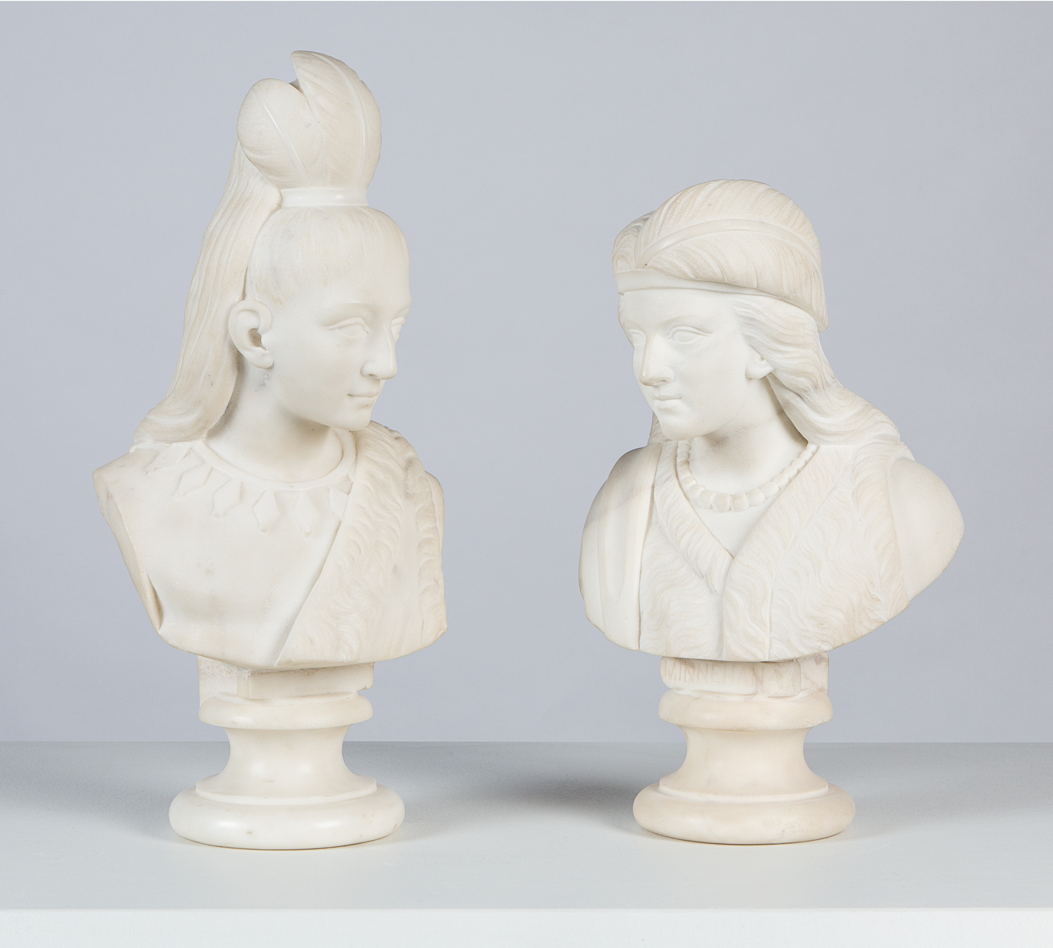 Two white marble statues facing each other. One wears their hair in a high ponytail adorned with feathers, the other wears their hair down with one large feather laying on one side of their head.