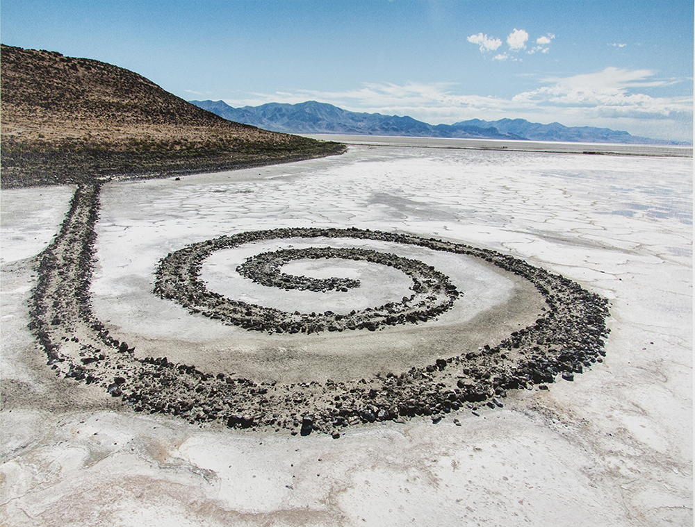 a photograph of an aerial view of a long jetty with a spiral at the end of the jetty. The jetty extends out into a dry salty lakebed with pink water at the edges of the photograph