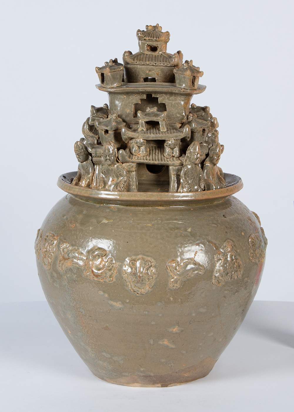 a gray-green urn with a squat round body and carved figures on the lid