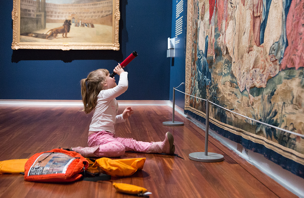 a little girl about four years old sits on the floor in front of a large European tapestry, she looks up at it through an orange toy telescope 